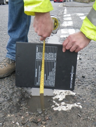 A man in work boots and a high vis jacket measuring a pothole with a tape measure.