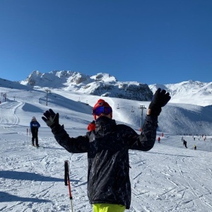 Man in ski equipment waving at the camera whilst stood in the snowy mountains.