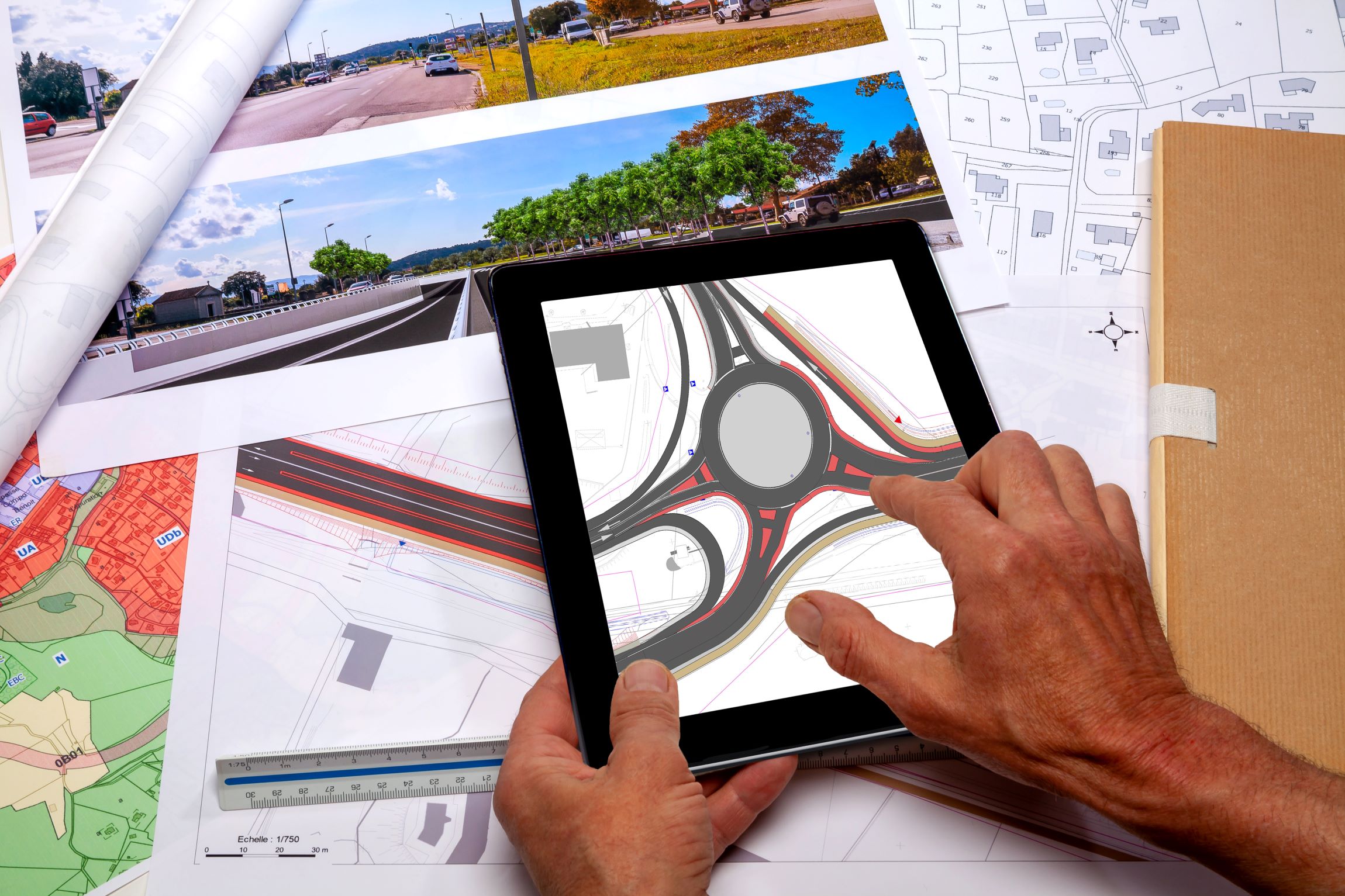Town planning - Land use planning - Employee holding in his hands a digital tablet displaying a road project plan, above a desk where ground plan maps are placed