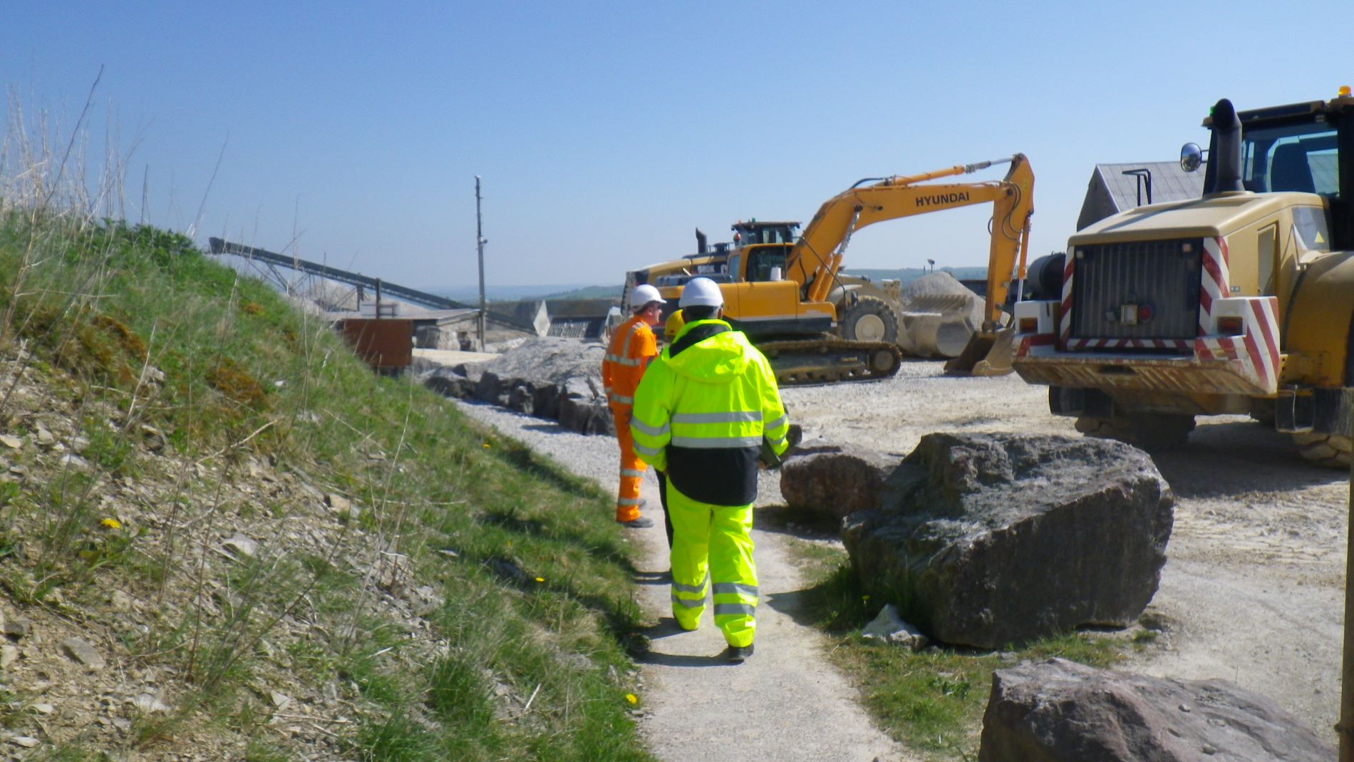 A group of people wearing high vis outfits stood next to large excavators, on the site of a quarry.