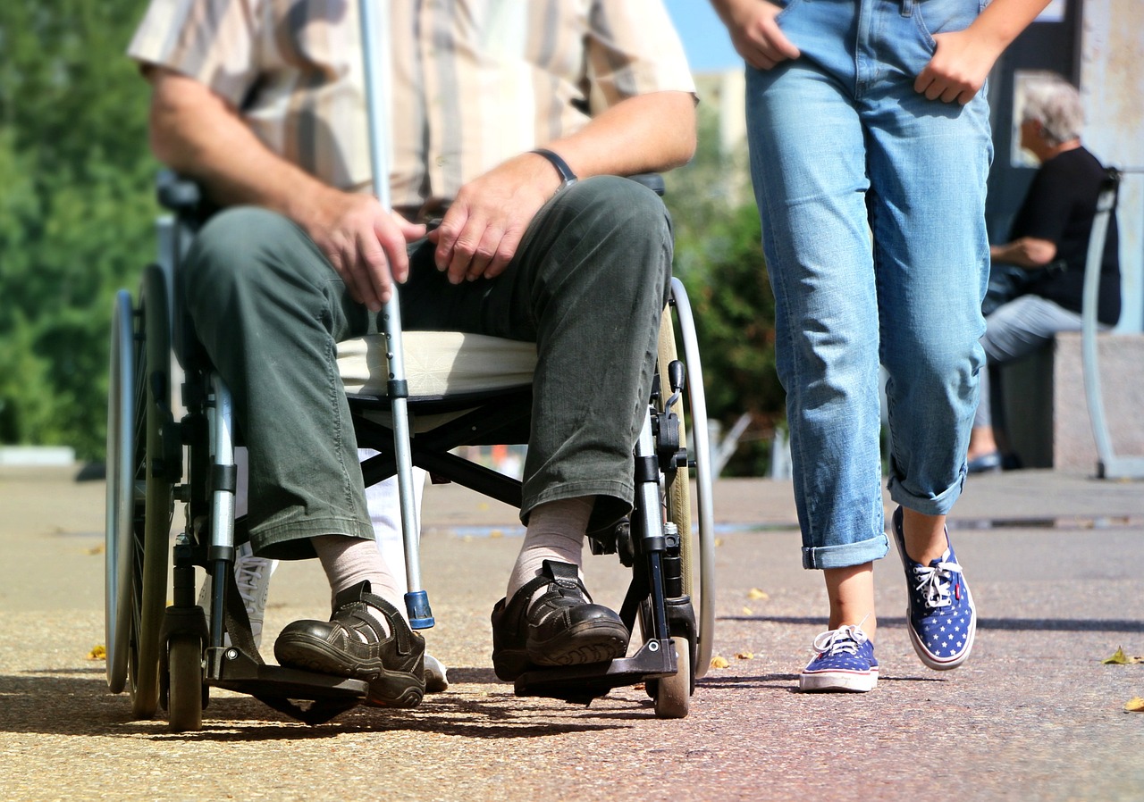 Man using a wheelchair holding a walking stick with someone walking next to him