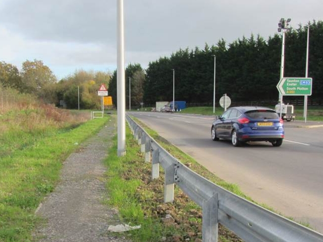 Narrow footpath alongside a dual carriageway with metal Armco barrier, road signs directing to Taunton, Exeter and South Molton via the M5, with overgrown verge and trees in the background