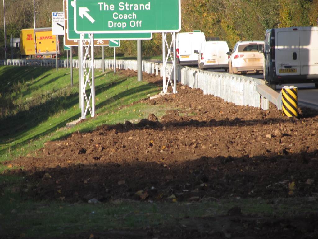 Roadside construction area beside a busy road showing disturbed ground and Armco safety barrier, with a green directional sign for The Strand Coach Drop Off and a DHL lorry visible in traffic