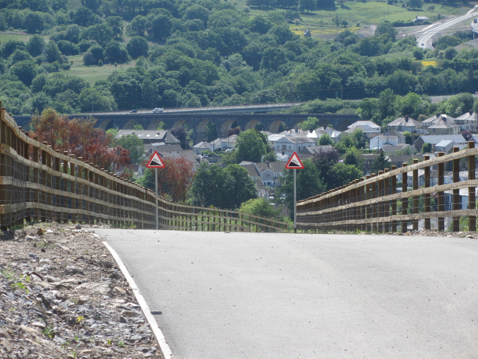 Newly constructed road descending a steep gradient with timber post-and-rail fencing on both sides, 10% gradient warning signs, and a panoramic view of a Welsh valley town with a stone viaduct and wooded hillside beyond