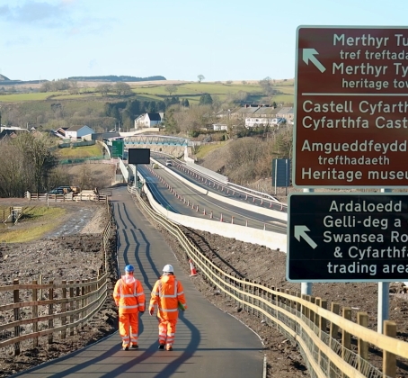 Two highway engineers in orange high-visibility PPE walking along a newly completed shared-use path at a road construction site near Merthyr Tydfil, Wales, with bilingual directional signs pointing to Cyfarthfa Castle, heritage museums and local trading areas.