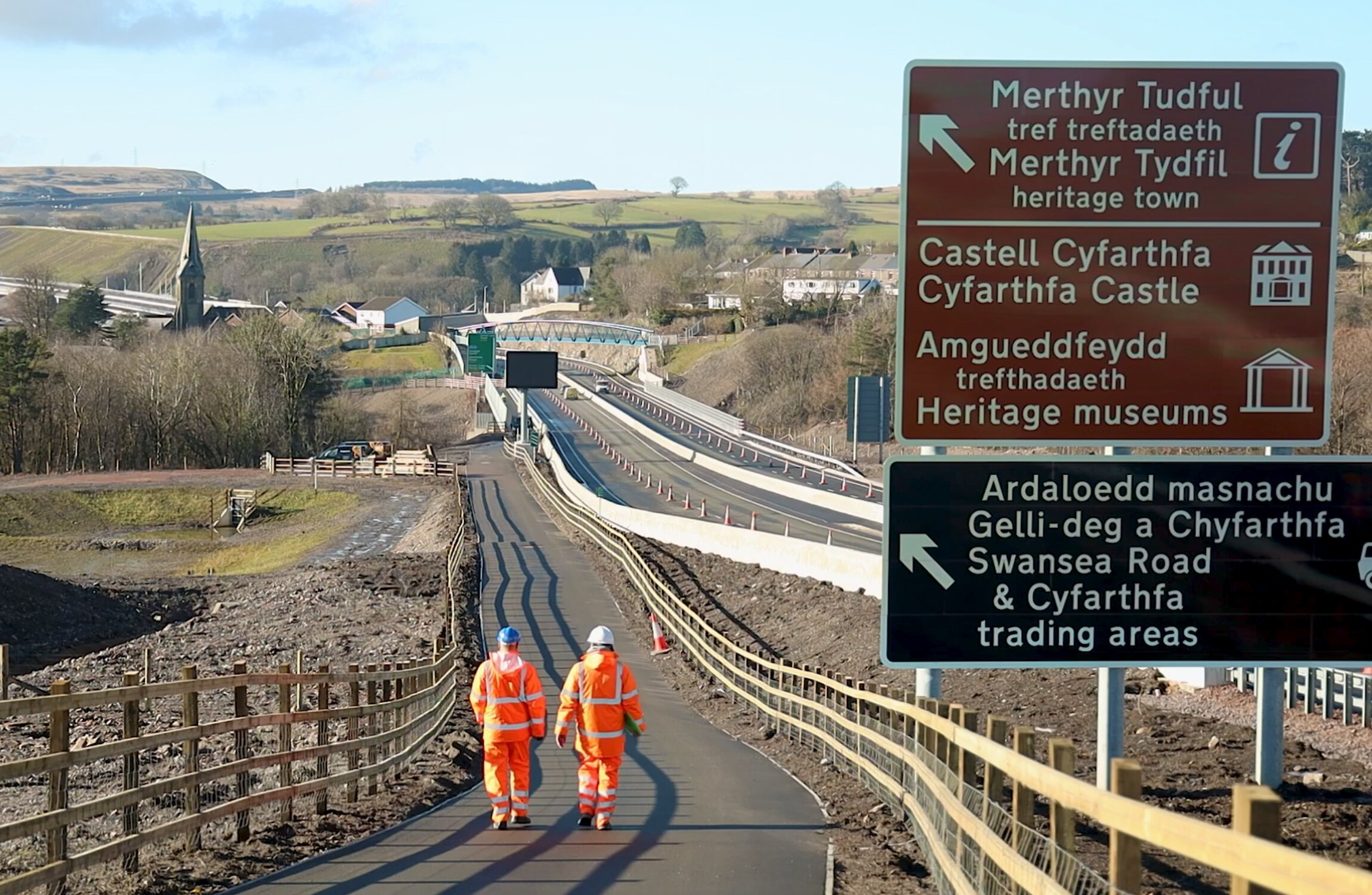 Two highway engineers in orange high-visibility PPE walking along a newly completed shared-use path at a road construction site near Merthyr Tydfil, Wales, with bilingual directional signs pointing to Cyfarthfa Castle, heritage museums and local trading areas.