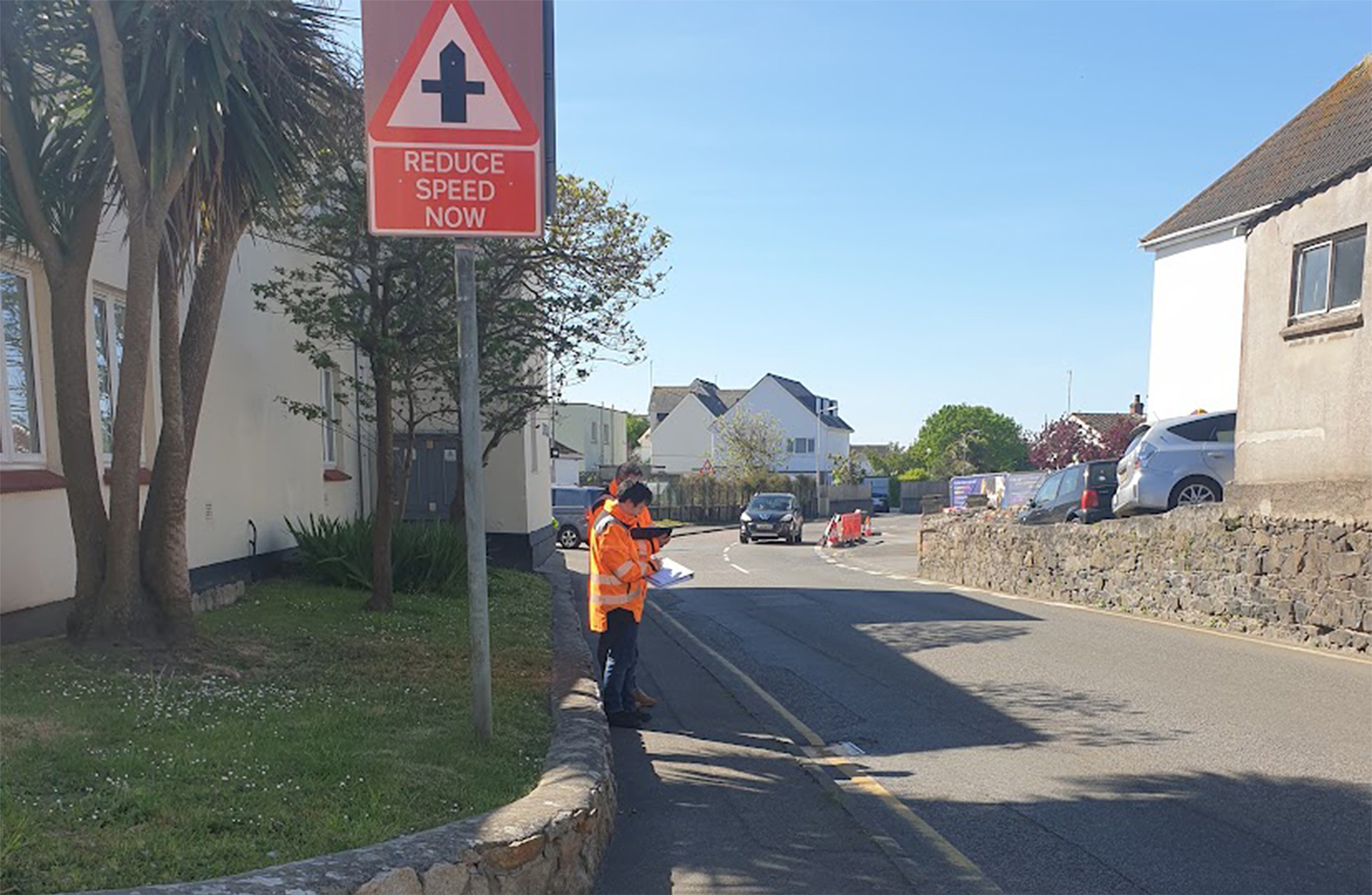A road in Jersey with two people standing on the walkway with two high visibility jacket.